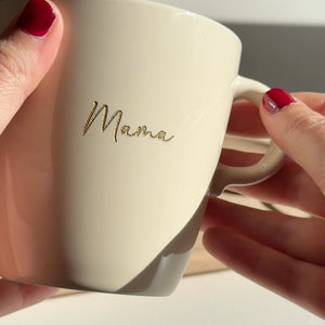 Twee handen houden een beige mok met de gegraveerde naam Mama in gouden letters vast in het zonlicht