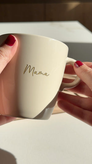 Twee handen houden een beige mok met de gegraveerde naam Mama in gouden letters vast in het zonlicht