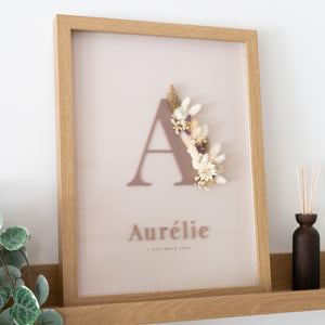 Letterkader met initiaal en naam in kokoskleurige acryl op een ivoren achtergrond, met droogbloemen in bruin en crème