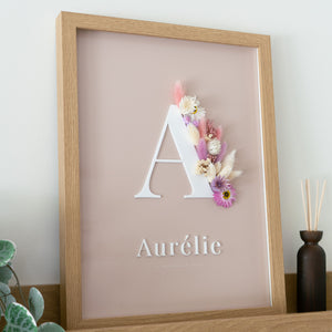 Letterkader met initiaal en naam in witte acryl op een nude achtergrond, met droogbloemen in roze en crème, op een houten schilderplank