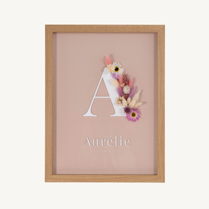 Vooraanzicht van een letterkader met initiaal en naam in witte acryl op een nude achtergrond, met droogbloemen in roze en crème