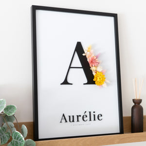 Letterkader met initiaal en naam in zwarte acryl op een witte achtergrond, met droogbloemen in geel en roze