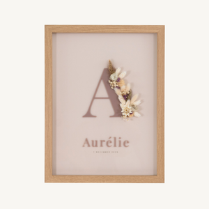 Vooraanzicht van een letterkader met initiaal en naam in kokoskleurige acryl op een ivoren achtergrond, met droogbloemen in bruin en crème