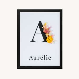 Vooraanzicht van een letterkader met initiaal en naam in zwarte acryl op een witte achtergrond, met droogbloemen in geel en roze
